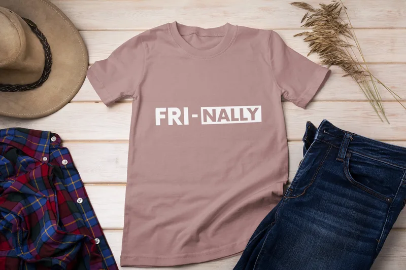 Light pink t-shirt with FRI-NALLY printed in white, laid out on a wooden surface