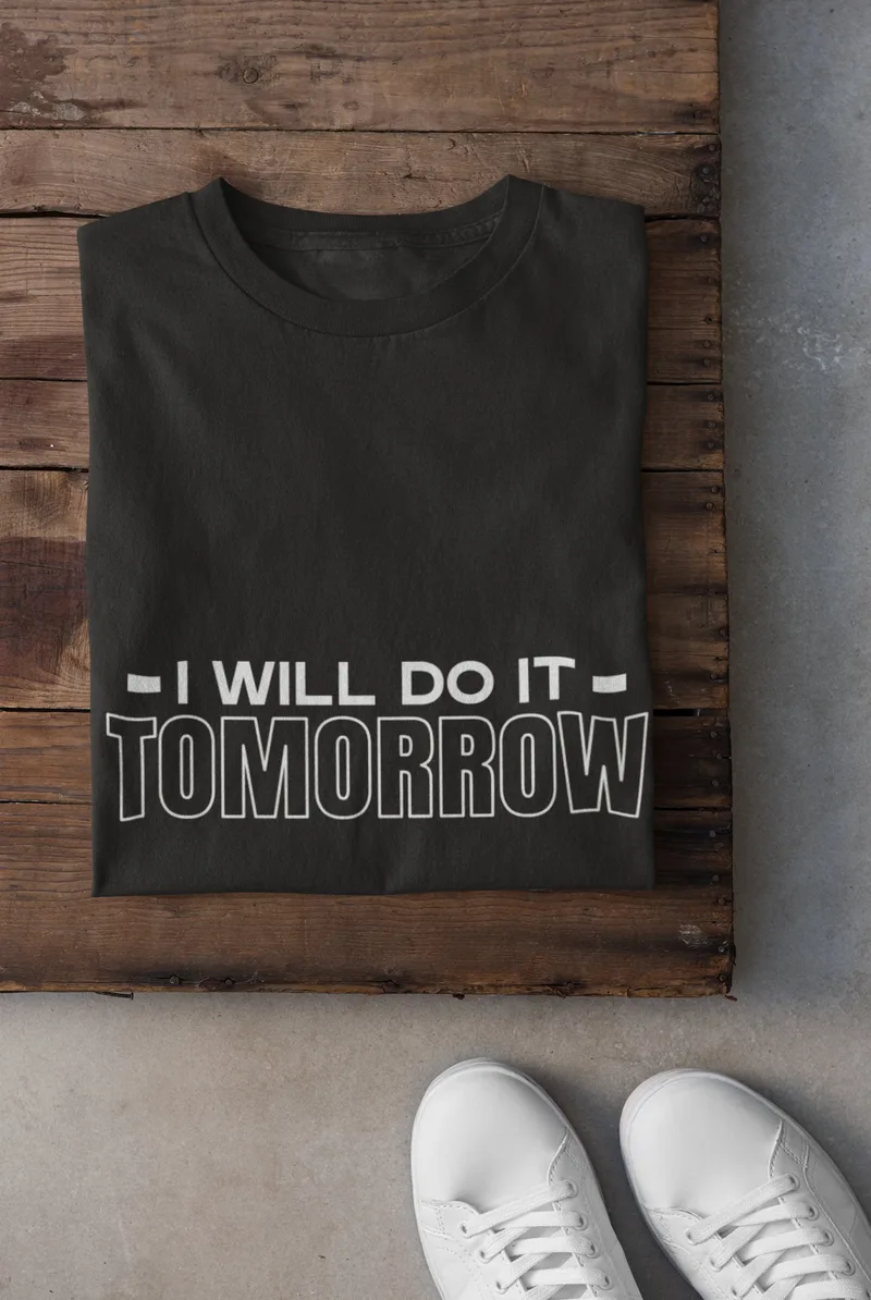 Black t-shirt with white text reading I will do it tomorrow, folded on a wooden surface