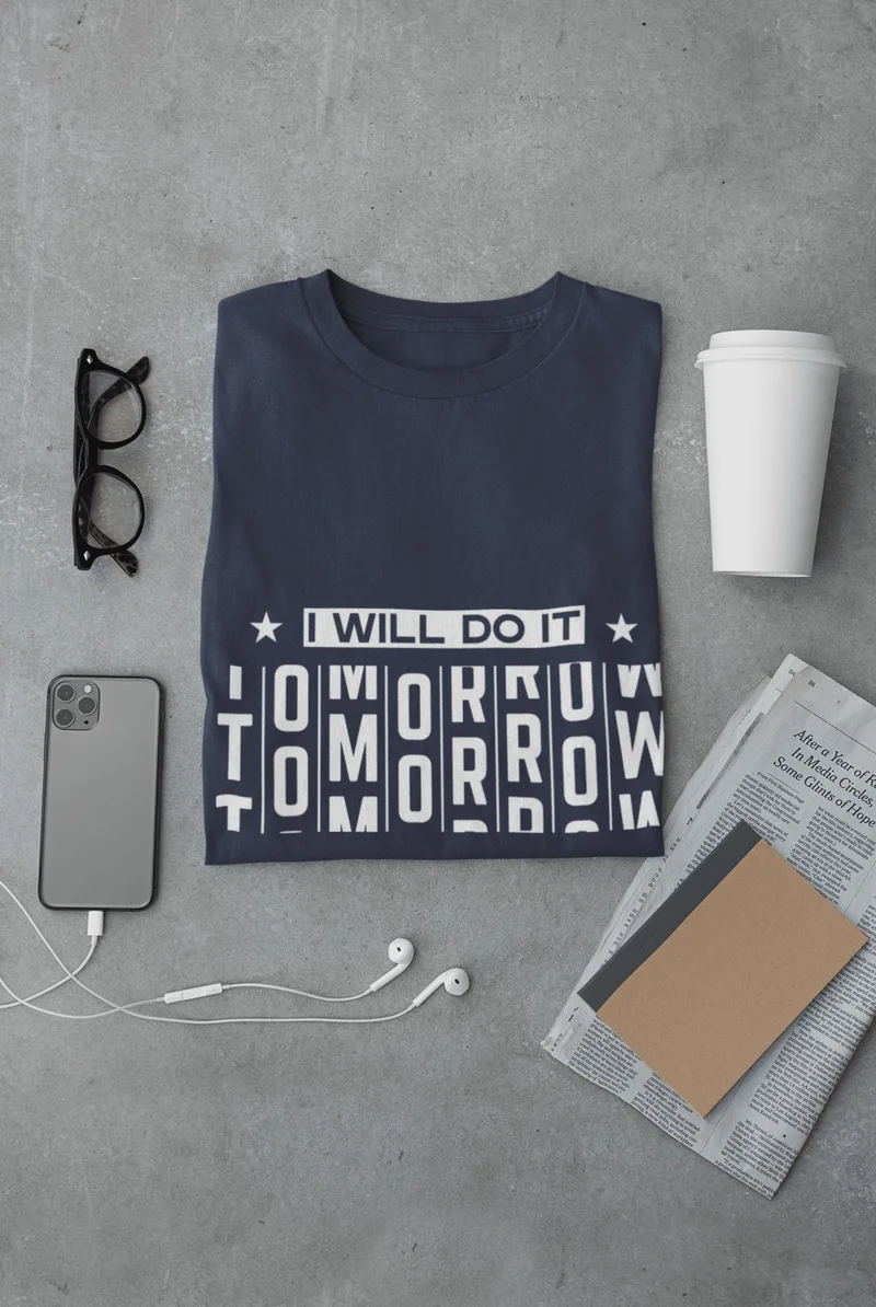 Navy blue t-shirt with I WILL DO IT TOMORROW printed in white, laid flat beside glasses and a notebook